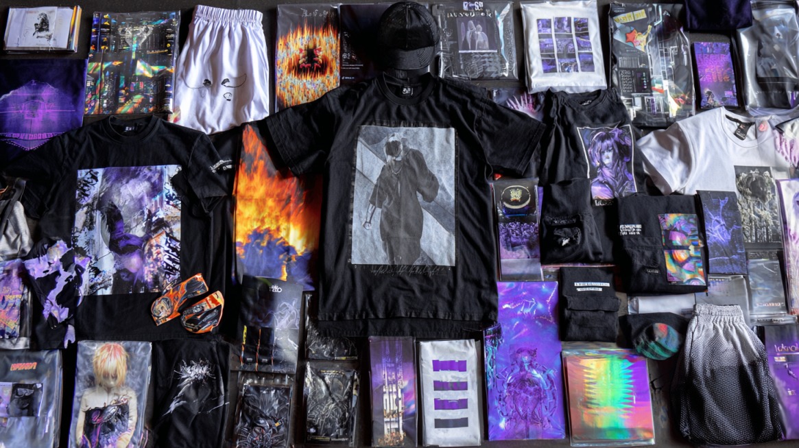 Flat lay of a diverse artist merch collection including graphic t-shirts, hoodies, shorts, posters, lanyards, caps, and holographic packaged items in a cohesive black, purple, and neon color palette